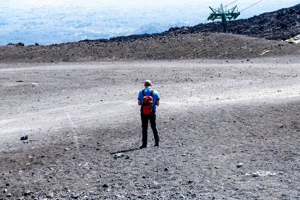 Catania, Sizilien - İtalya - 04-22-2025: Kırmızı sırt çantalı bir kişi Etna Dağı 'nın gri volkanik ovasında duruyor, seyrek kayalar ve uzak teleferik yapısıyla çevrili..