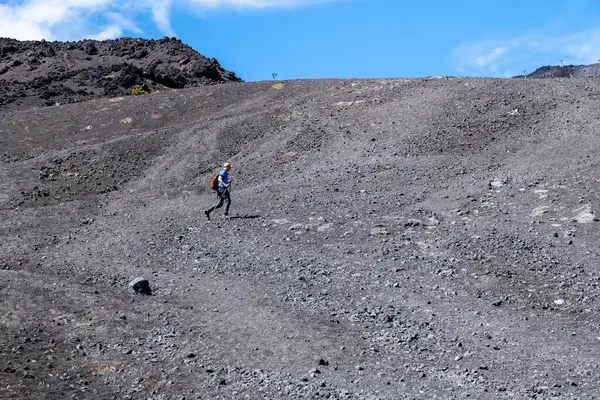 Catania, Sizilien - İtalya - 04-22-2025: Kırmızı sırt çantalı bir yürüyüşçü görünür lav kayaları ve sırtları olan parlak mavi gökyüzü altında, geniş, gri bir volkanik yamaçta yokuş yukarı yürüyor.