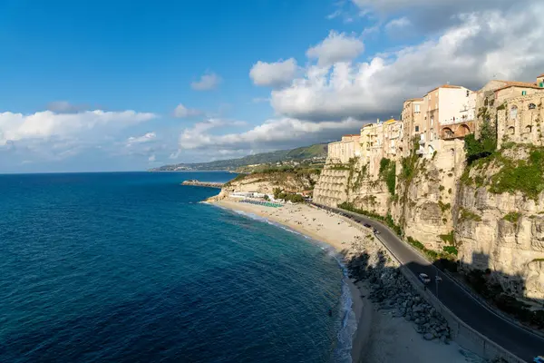 Tropea - Kalabrien - İtalya - 05-29-2025: Calabria 'daki Tropea, kumlu bir sahil üzerindeki tarihi uçurumlar ve parçalı bulutlu bir gökyüzünün altındaki açık mavi deniz üzerindeki tarihi binaları gösteriyor.