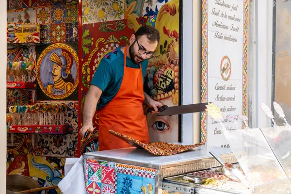 Pizzo - Kalabrien - İtalya - 06-02-2025: Turuncu önlüklü bir adam Pizzo, Calabria, İtalya 'da canlı dekore edilmiş bir sokak tezgahında büyük renkli bir nugatı dilimliyor.