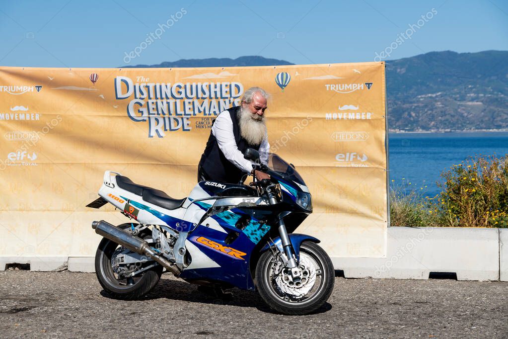 Reggio Calabria - Kalabrien - 05-18-2025: Man with long beard next to Suzuki GSX-R during the Distinguished Gentlemans Ride in Reggio Calabria with banner and coast in background.