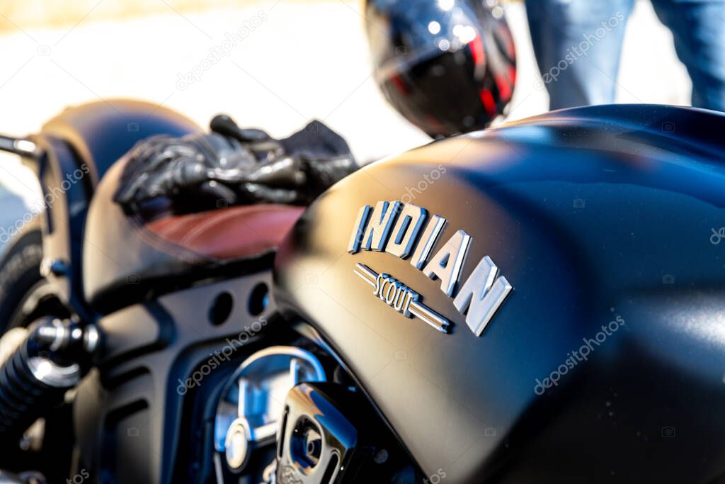 Reggio Calabria - Kalabrien - 05-18-2025: Close-up of a matte black Indian Scout motorcycle showing tank logo, seat with gloves, and blurred background with partial helmet and legs.
