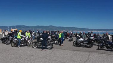 Motosikletliler seçkin beyefendiler için toplanıyor. Kameralar sağa dönerken, park halindeki bisikletleri, denizi ve yoldan geçen gümüş bir arabayı gösteriyor..