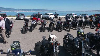 Reggio Calabria 'da bir sahil toplantısında sağdan sola park etmiş motorsikletler gösteriliyor. İnsanlar motosikletlere hayran kalıyor ve mavi gökyüzünün altında konuşuyor..