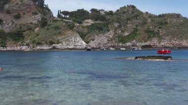 Isola Bella, Taormina, Sicilya 'da kalabalık bir plaj sahnesi, kameralar kıyı şeridinde yavaşça hareket ederken insanların dinlendiğini ve yürüdüğünü gösteriyor..