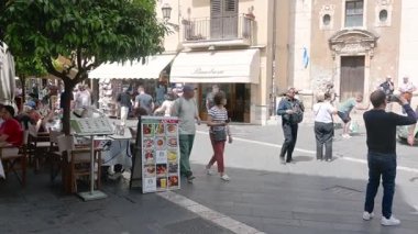 Turistler ve yerel halk Taormina, Sicilya 'da tarihi bir meydanda yürüyor. Arka planda klasik bir araba ve dükkan önleri görünüyor..