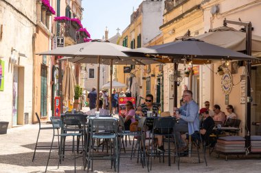 Matera - Basilikata - Italien - 06-16-2025: İnsanlar Matera, İtalya 'nın dar bir caddesinde güneşli bir günde, eski binalar ve canlı çiçeklerle çevrili açık bir kafede oturuyorlar.