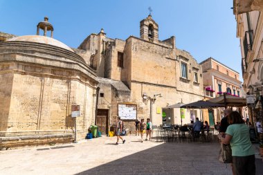 Matera - Basilikata - Italien - 06-16-2025: İtalya 'nın Matera kentinde tarihi kireçtaşı binalar ve şemsiyeli bir açık hava kafe ile çevrili güneşli bir meydanda yürüyen turistler.