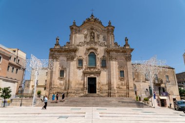 Matera - Basilikata - Italien - 06-16-2025: Matera, İtalya 'da Chiesa di San Francesco dAssisi' nin Barok cephesi, mavi gökyüzünün altında basamaklarda ve kare şeklinde yürüyen turistlerle.