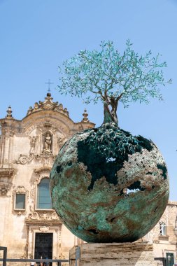 Matera - Basilikata - Italien - 06-16-2025: İtalya 'nın güneyindeki Matera kentinde bir barok kilisesinde sergilenen, üzerinde açık kökleri ve zeytin ağacı bulunan bir kürenin bronz heykeli.