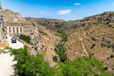 Matera - Basilikata - Italien - 06-16-2025: View of Matera in Basilicata with historic buildings overlooking a rocky canyon, green valley and hiking paths under clear summer sky.