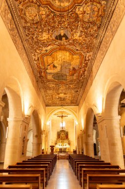 Matera - Basilikata - Italien - 06-16-2025: Matera 'daki Chiesa di San Pietro Caveoso' nun içi boyalı ahşap tavanı, kemerli nav, banklar ve yanan sunağın üzerinde haç.
