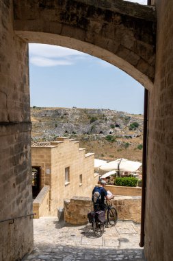Matera - Basilikata - Italien - 06-16-2025: Matera, Basilicata 'da kemerin altında bisikletli bir adam, mağaralara bakan kanyon, taş binalar, şemsiyeler ve restoran terası olan bir adam.