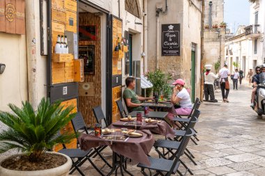 Matera - Basilikata - Italien - 06-16-2025: Matera 'da insanların tarihi binalar ve kaldırımlarla çevrili kırsal bir restoranın açık masalarında yemek yediği sokak sahnesi.