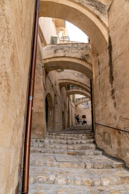 Matera - Basilikata - Italien - 06-16-2025: Matera, Basilicata 'da yüksek kemerli dar tarihi dar bir sokakta taş merdiven, üzerinde bisiklet ve köpek olan bir kişiyi gösteriyor.