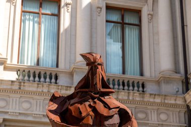 Tarihi neoklasik sütunları olan tarihi bir binanın önünde sergilenen, miğferli bir figürü andıran paslı bir metal heykelin yakın çekimi.