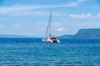 Scilla, Calabria kıyılarına demirlemiş katamaran, dağınık şamandıralar ve uzak yeşil dağ manzarasıyla parçalı bulutlu bir gökyüzü altında.