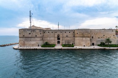 Simetrik kuleleri, İtalyan bayrağı, palmiye ağaçları ve bulutlu gökyüzünün altında deniz kenarında taş gezinti yapan tarihi kıyı kalesinin ön manzarası..