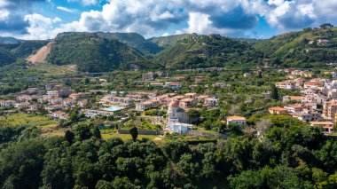 Calabria 'daki Tropea' nın hava manzarasında dağınık evler, kubbeli bir kilise, yeşil tepeler, yoğun bitki örtüsü ve dramatik bulutlu gökyüzü görülüyor..