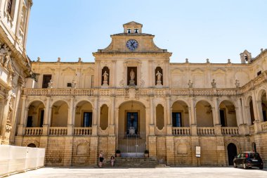 Lecce - Lecce - İtalya - 06-21-2025: Merkezi cephede kemerli yürüyüş yolları, korkuluklar, heykeller ve girişin üzerinde tanınmış bir saat yer alıyor. Lecce - İtalya konumu.