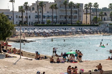 Otranto - Apulien - İtalya - 06-20-2025: İtalya 'nın Otranto kentinde güneş banyosu yapanlar, yüzücüler, şemsiyeler ve apartmanların yer aldığı bir halk sahnesi.