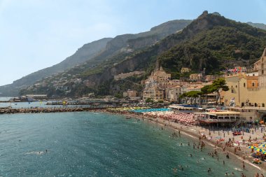 Amalfi - Kampanien - Italien - 07-04-2025: Şemsiyeli ve banyolu kalabalık plaj, açık mavi su, liman binaları, dik tepeler ve arka planda kıyı yolu. Stadt Amalfi