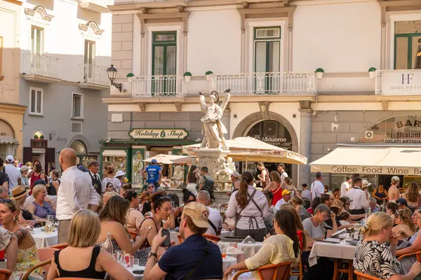 Amalfi - Kampanien - Italien - 07-04-2025: Amalfi kentindeki açık hava restoranında bir heykel ve gün ışığında tarihi binaların yakınlarındaki masalarda yemek ve içmek.