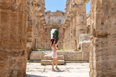 Capaccio Paestum - Kampanien - Italien - 07-06-2025: Başörtüsü takan dövmeli bir kadın güney İtalya 'da bulunan Paestum' daki bir tapınağın içindeki antik kalıntıların fotoğrafını çekiyor.