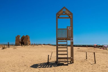 Cerveteri - Latium - Italien - 07-30-2025: İnsanlar Torre Flavia 'nın antik taş kalıntılarının yakınında şemsiyeler altında dinlenirken sahilde tahta bir cankurtaran kulesi duruyor.