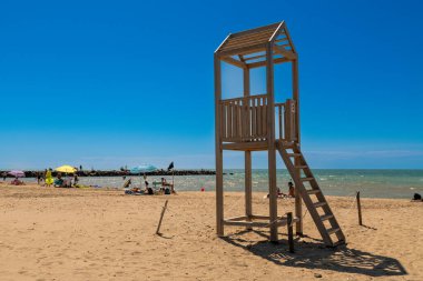 Cerveteri - Latium - Italien - 07-30-2025: Suya yakın kumlu bir sahilde duran ahşap bir cankurtaran kulesi, güneşlenenler, şemsiyeler ve canlı mavi bir yaz gökyüzü ile çevrili.