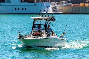 Civitavecchia - Latium - Italien - 07-31-2025: Balıkçı çubukları ve ekipmanlarıyla küçük beyaz bir motorlu teknede bulunan iki adam, daha büyük rıhtımlı tekneleri olan bir marina yakınındaki sakin turkuaz sularda geziniyorlar..