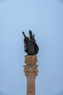 İtalya 'nın Lecce şehrinde süslü bir sütunun üzerinde duran, üzerinde Mitre ve Crosier olan cüppeli bir piskopos heykeli, açık mavi bir gökyüzüne karşı fotoğraflandı..
