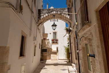 Locorotondo 'da, bembeyaz yıkanmış taş binalar, kemerli yürüyüş yolları, fenerler ve açık mavi gökyüzünün altında balkon olan dar bir sokak..