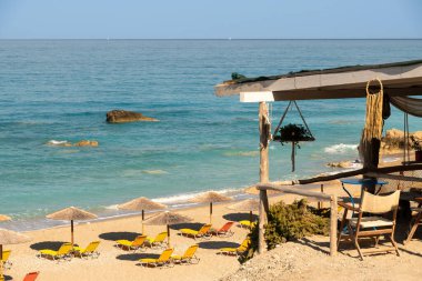Turkuaz denizin yanındaki kumlu sahilde saman şemsiyeli sarı ve turuncu plaj sandalyeleri kırsal ahşap bir yapıdan görülüyordu..