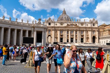 Roma - Lazio - İtalya - 08-02-2025: Büyük bir grup insan parçalı bulutlu bir gökyüzü altında sütunlar, heykeller ve kubbelerle St. Peters Basilikac binasının önünde yürüyor.