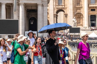 Roma - Lazio - İtalya - 08-02-2025: Aralarında mavi şemsiyeli bir papazın da bulunduğu bir grup insan, St. Peters Meydanı 'ndaki tarihi bir taş binanın önünde parlak güneşin altında yürüyor.
