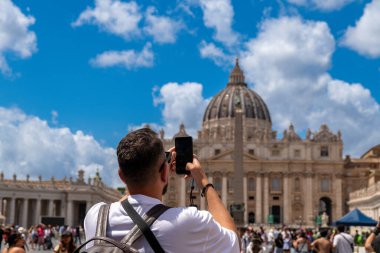 Roma - Lazio - İtalya - 08-02-2025: Bir adam St. Peters Bazilikası 'nı turistler arasında hareketli mavi gökyüzünün altında dururken akıllı telefon kullanarak fotoğraflıyor.