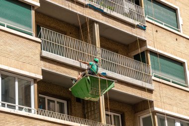 Roma - Lazio - İtalya - 08-02-2025: İşçi, balkonları, korkulukları ve kapalı panjurları olan bir binanın ön cephesine halatlarla bağlanmış bir konteynırda askıya alındı.