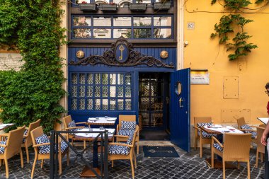 Roma - Lazio - İtalya - 08-02-2025: Modelleşmiş mavi girişi, hasır sandalyeleri ve beyaz masa örtüleri ve çatal bıçakları olan açık hava masaları olan bir restoranın dış görünüşü.