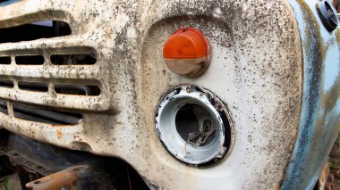Ağır yıpranmış ve paslanmış bir aracın ön tarafında, kayıp bir far ve bozulmamış turuncu bir gösterge ışığı görülüyor..