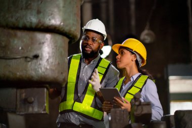 Afrikalı Amerikalı fabrika kadını tablet tutuyor ve iş arkadaşının makine işlevini ve bakım ya da onarım sürecini açıklamak için kullanıyor..