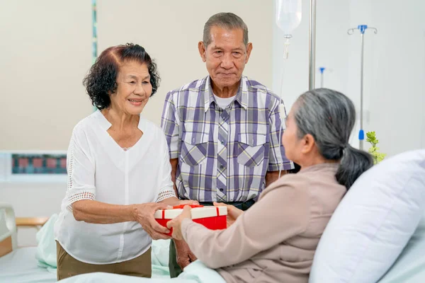 Asian senior woman stand with her husband and give the gift box or present to other senior woman to express and encourage during treatment in hospital.