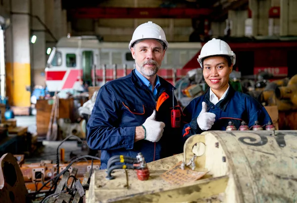 Close up and portrait of Caucasian worker man and Asian woman show thumbs up and look at camera with train in the background of repair and maintenance factory.