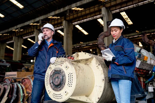 Asian factory woman concentrate with work using tablet and stand near big spare part of machine and her coworker use walkie talkie to discuss with the other worker in their team.