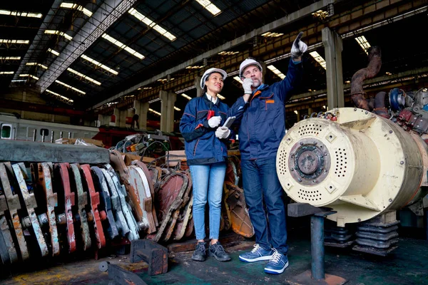 Wide shot of Caucasian factory man use walkie talkie to contact his team and stand with Asian co-worker woman near spare part of machine of the train in industrial business workplace.