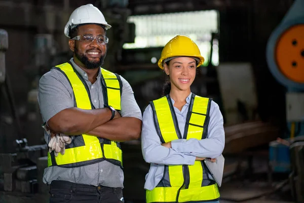 Couple of African American worker man and woman stand with arm crossed and also look to right in workplace area and some machine in the background.