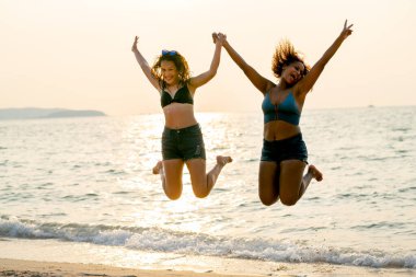 İki çaydanlık kızı tatilde ya da tatilde mutlulukla ve sıcacık ışıkla ileriye doğru zıplıyor ve arka plan olarak denize yansıyor..