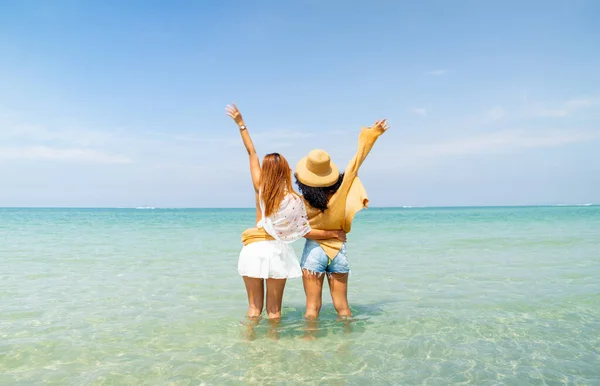 stock image Back of couple women stand with wave hands out to sea with happiness and enjoy holiday together at beach with day light and relax time.