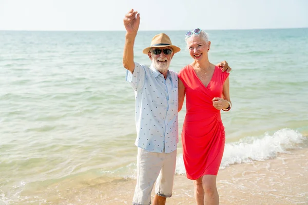 老人挥挥手 看着相机 也和妻子拥抱在一起 他们在度假或度假的海滩上放松的时候看起来很开心 — 图库照片