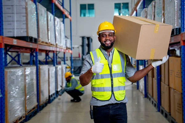 African American warehouse worker carry the box on his shoulder and show thumbs up to camera with smiling and his coworker check product on shelves in the background.